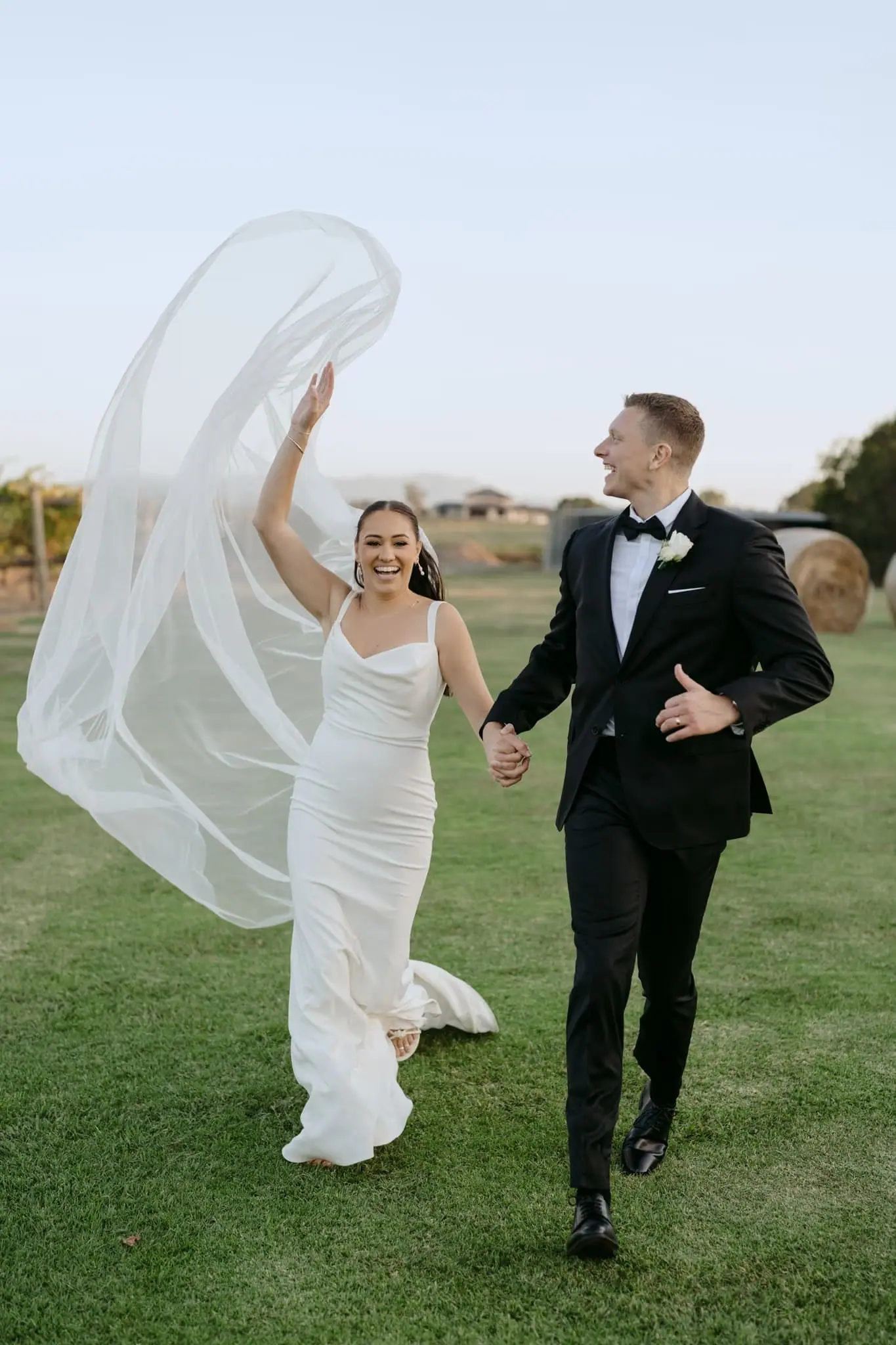Bride lifting her veil overhead while walking with the groom across a lawn.