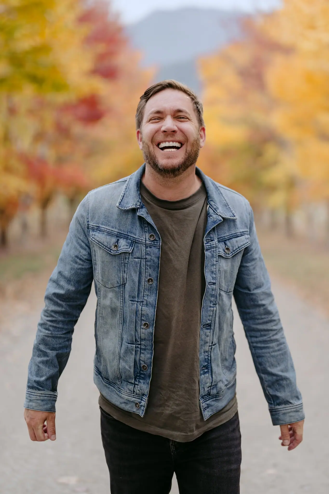 Man in a denim jacket laughing on an autumn tree-lined avenue.