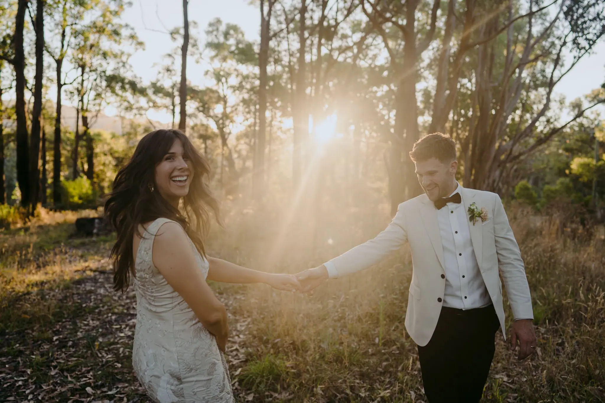 Bride and groom holding hands and laughing among sunlit forest trees.