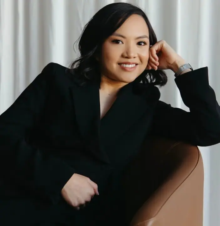 Woman in a dark suit smiling while seated beside curtains.