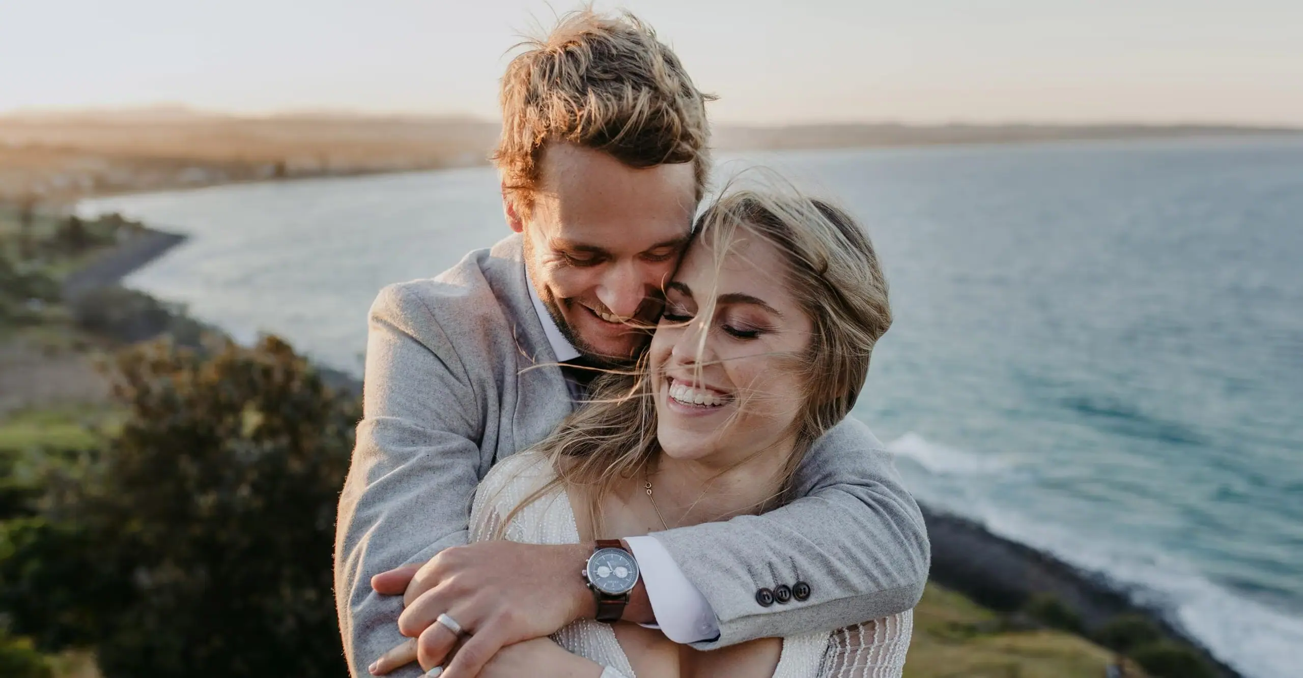 Groom embracing the smiling bride from behind beside coastal cliffs.