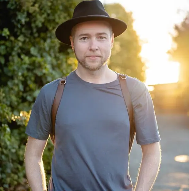 Man wearing a hat and camera straps standing beside a hedge at sunset.