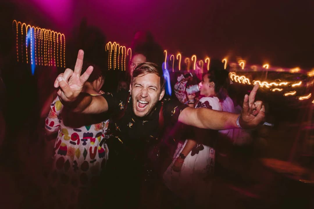 Man raising both hands and cheering beneath blurred colourful reception lights.
