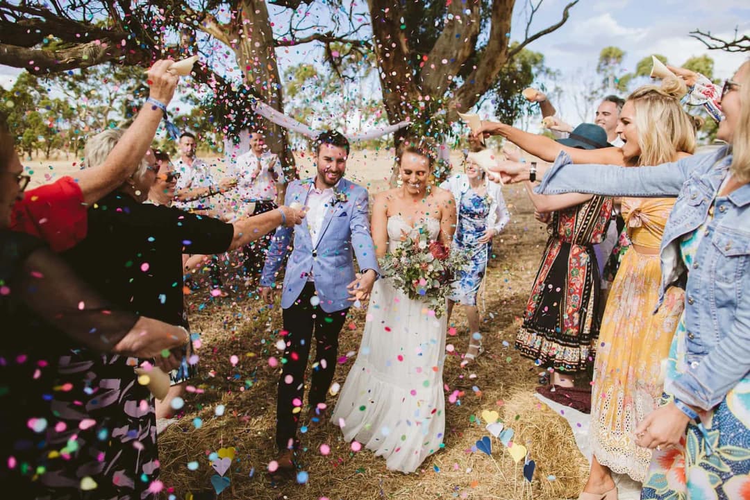 Couple walking beneath colourful confetti thrown by woodland ceremony guests.