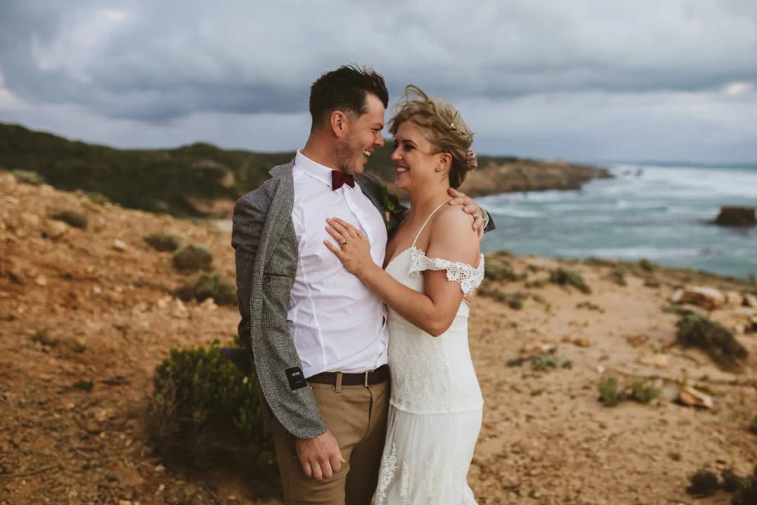 Bride and groom smiling at each other beside a rocky coastline.