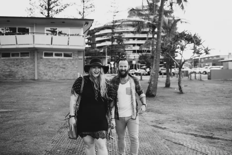 Black-and-white portrait of a couple walking beneath beachside pine trees beside apartment buildings.