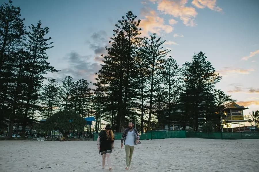 Couple walking across pale beach sand beneath tall pine trees and a cloudy sunset sky.