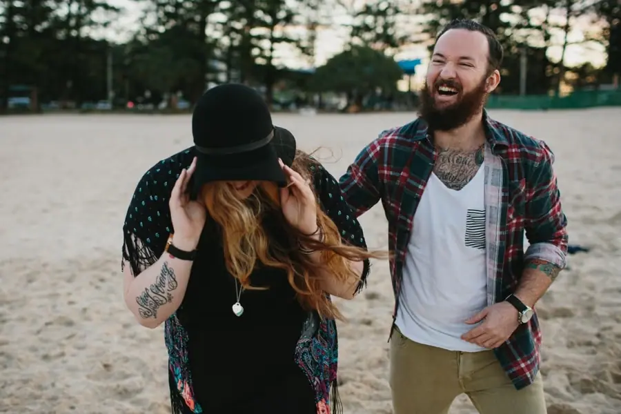 Bearded man laughing as the woman beside him holds the brim of her black hat on the beach.