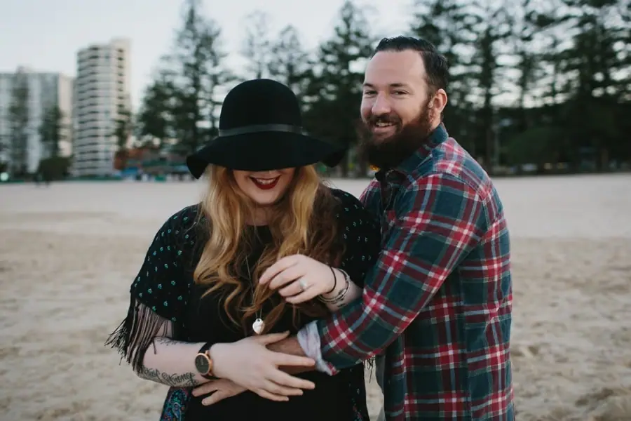 Bearded man wrapping his arms around his smiling partner in a black hat on the beach.