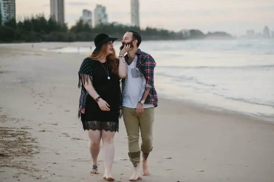 Couple walking barefoot along the shoreline, the woman reaching up toward her partner’s face.