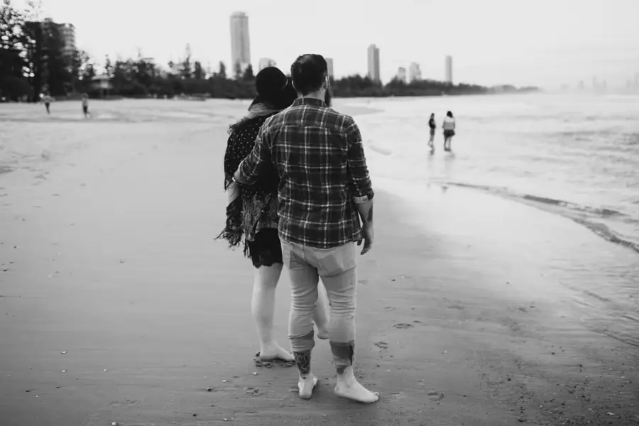 Black-and-white photograph of a couple standing together at the water’s edge, viewed from behind.