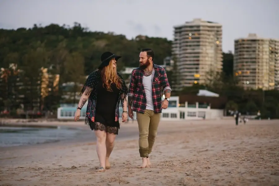 Couple walking barefoot together across the beach with trees and apartment towers behind them.