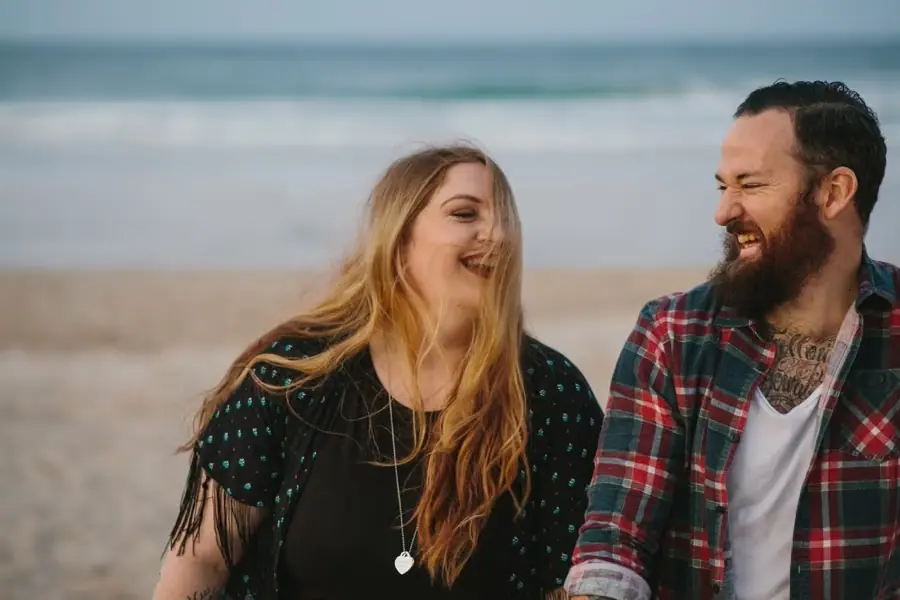 Woman laughing beside her smiling bearded partner with the sea behind them.