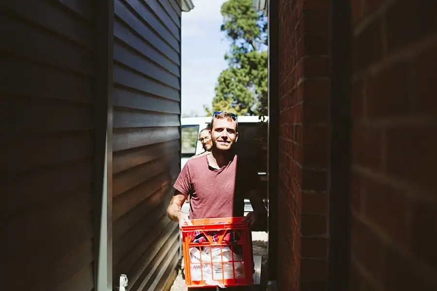 Man carries a red crate through a narrow outdoor passage.