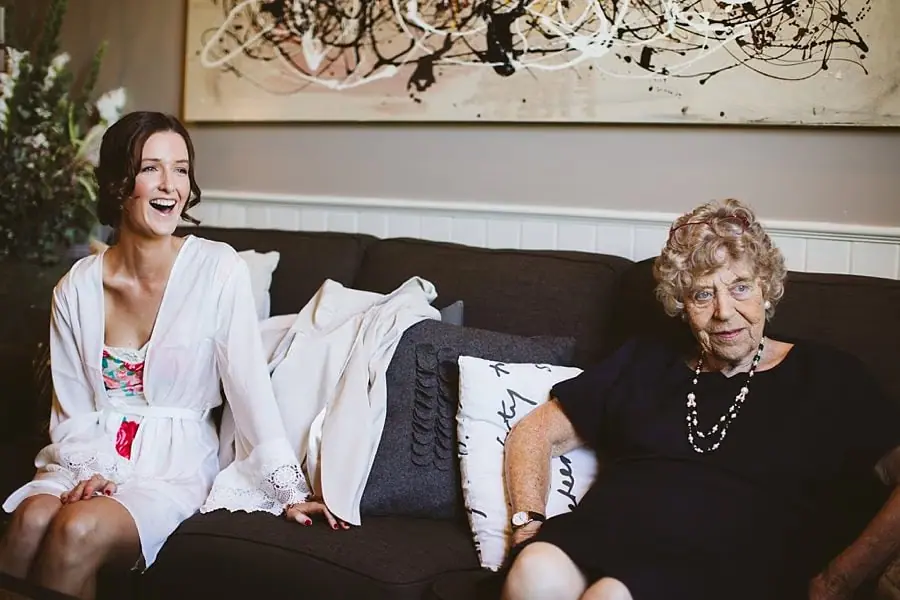 Bride in a robe laughs beside an older woman on a sofa.