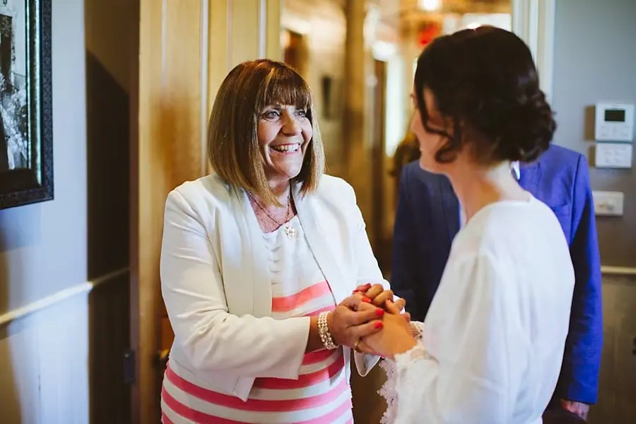 Bride smiles while holding an older woman's hands indoors.
