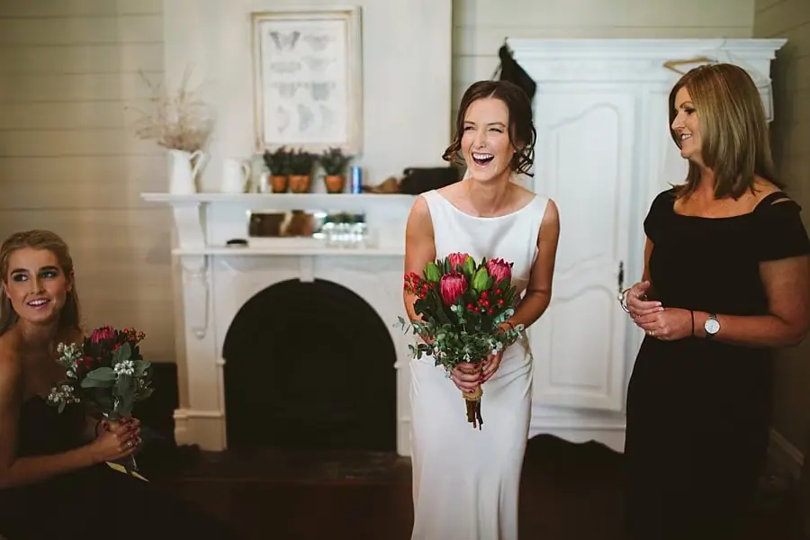Bride laughs while holding her bouquet beside friends indoors.