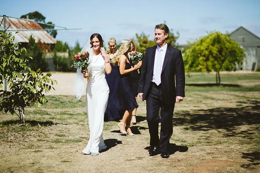 Bride approaches across the lawn with a suited man beside her.