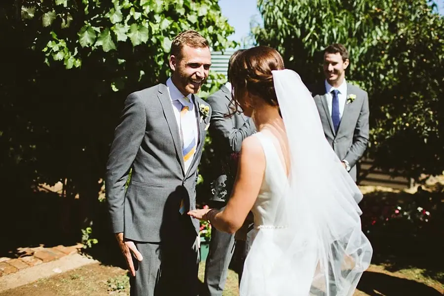 Groom smiles toward the bride beneath garden foliage.