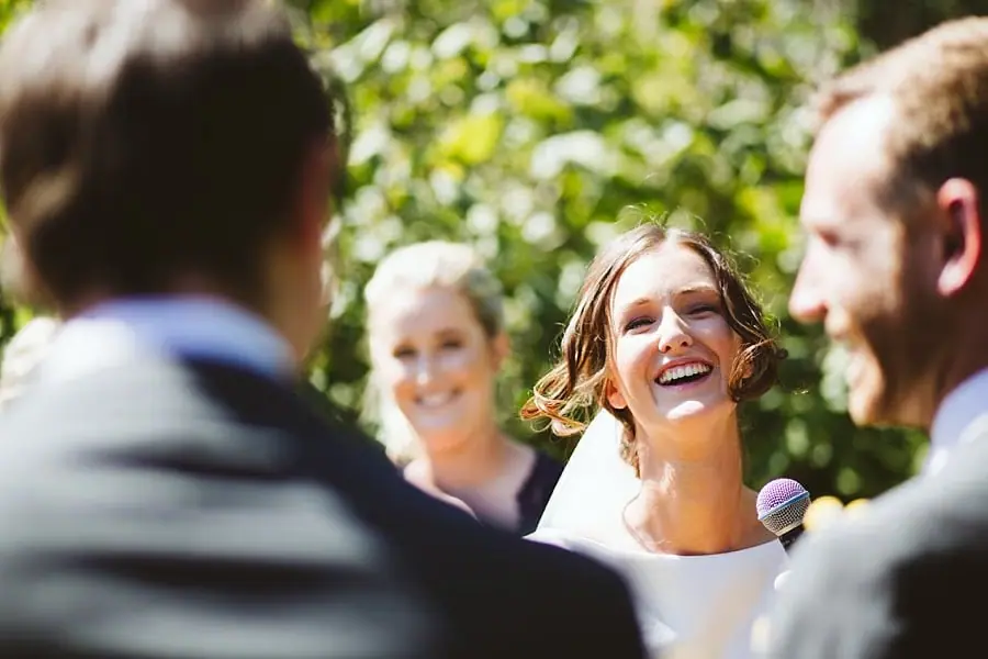 Bride laughs toward the groom while holding a microphone.