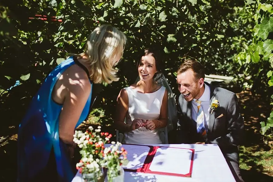 Couple laughs with a guest beside wedding paperwork in the garden.