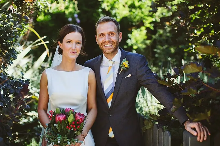 Couple poses beside leafy greenery with the bride holding a colourful bouquet.