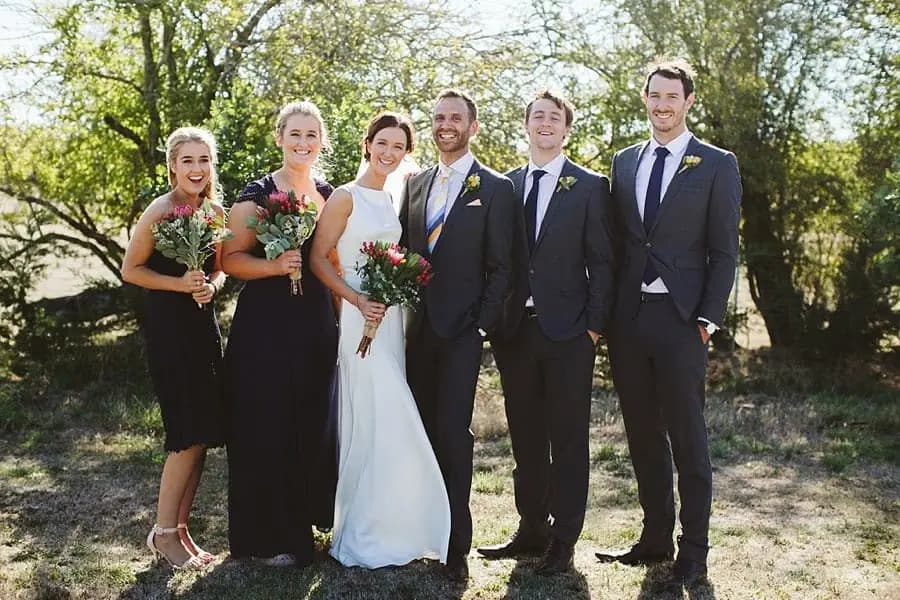 Wedding party poses beneath sunlit trees with bouquets.