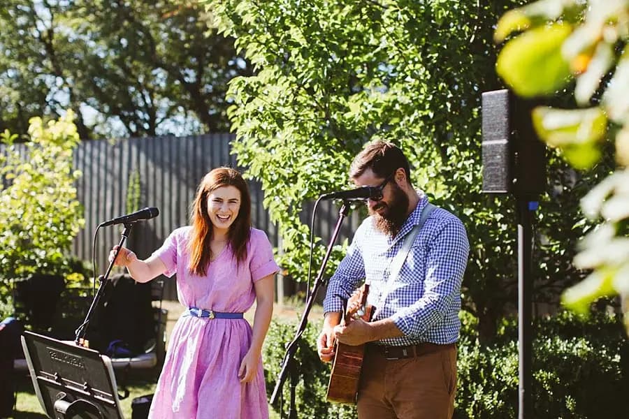 Singer and guitarist perform beneath leafy garden trees.