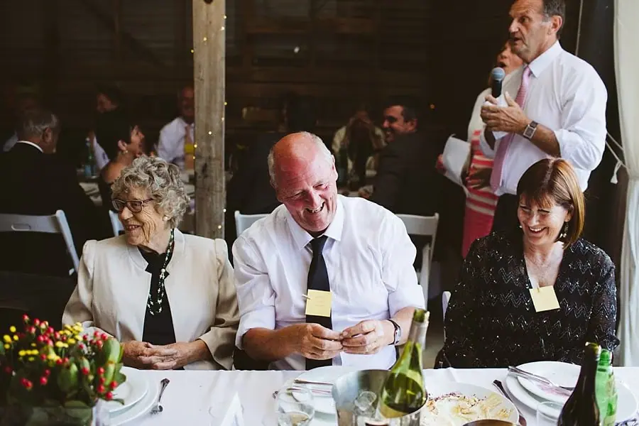 Guests laugh at their reception table while a man speaks into a microphone.