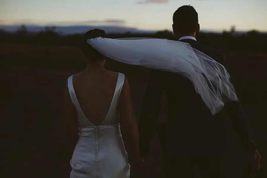 Rear view of the couple embracing as the bride's veil blows across them at dusk.