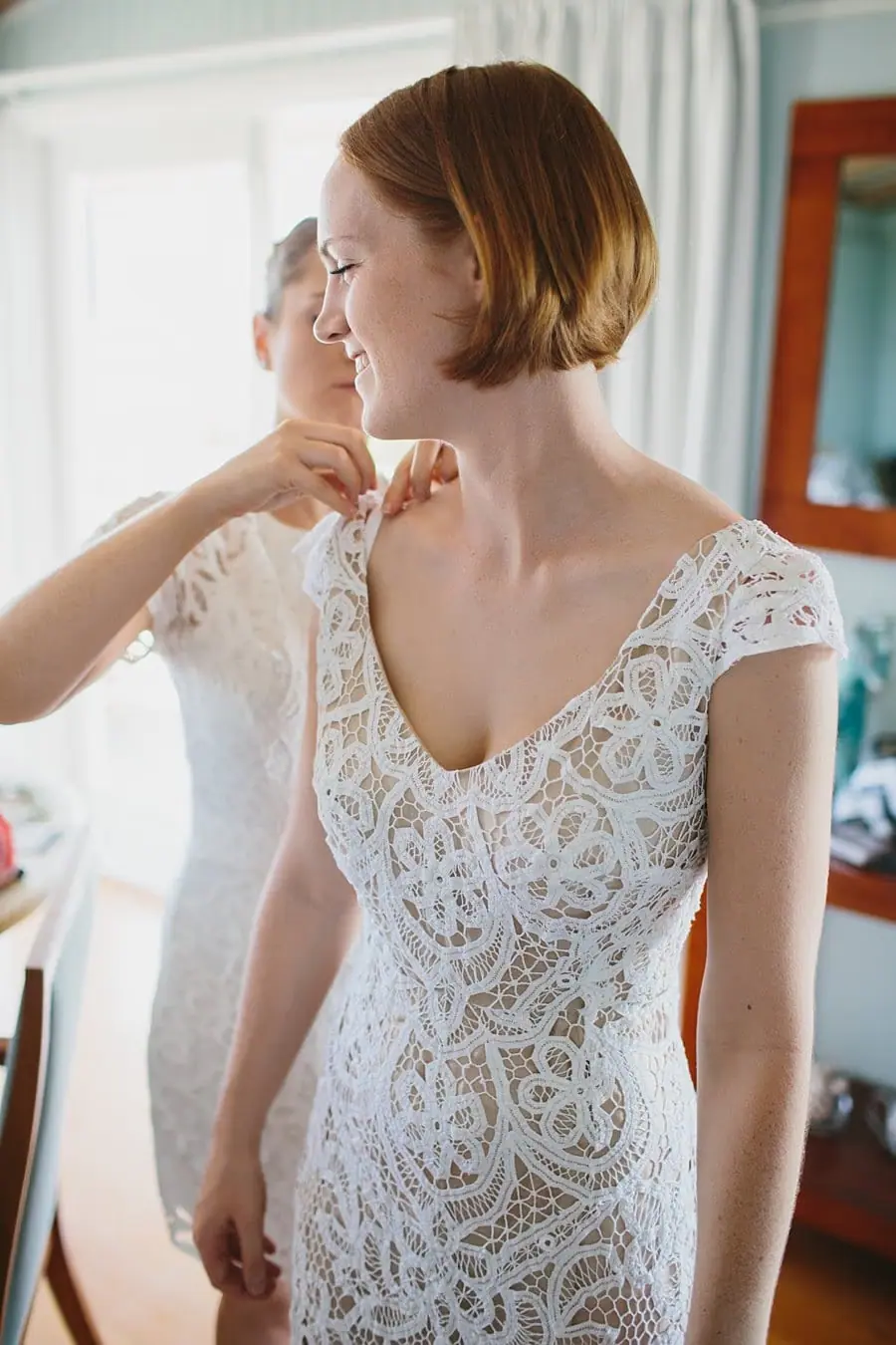 Bride standing indoors in a white lace dress while someone adjusts her shoulder.