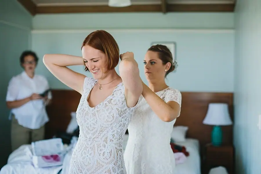 Bride raising her arms as a companion helps fasten her dress.