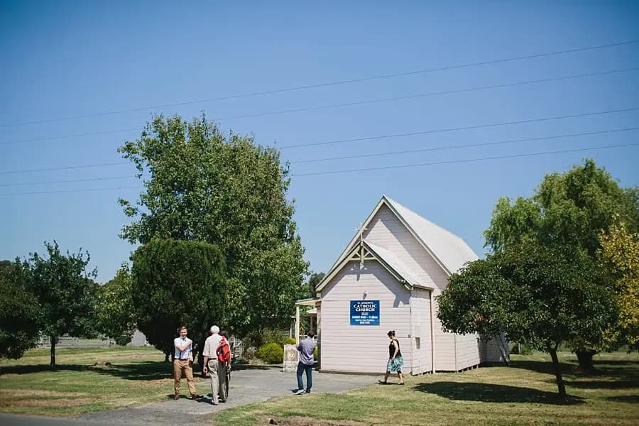 Guests gathered outside a small pale timber church beneath a blue sky.