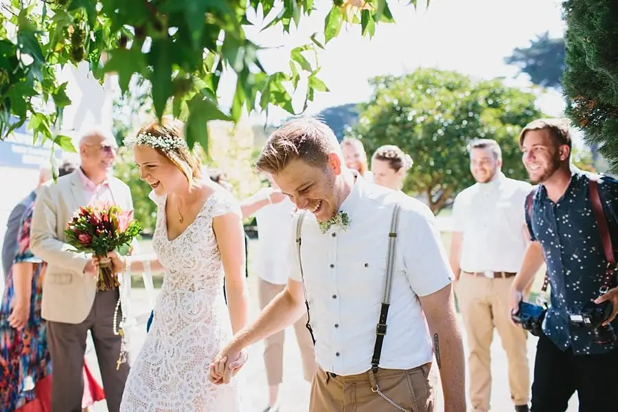 Bride and groom walking among guests beneath leafy trees.
