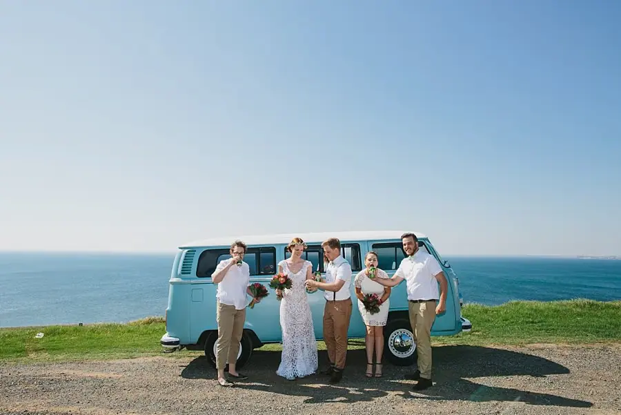 Wedding party posing beside a pale blue van on a grassy coastal lookout.