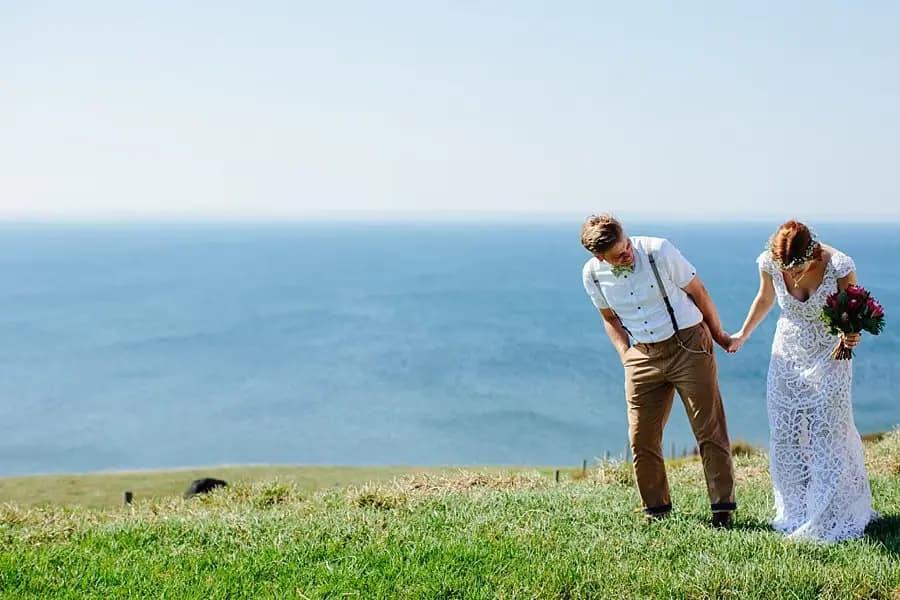 Bride and groom holding hands and looking down on a grassy coastal slope.
