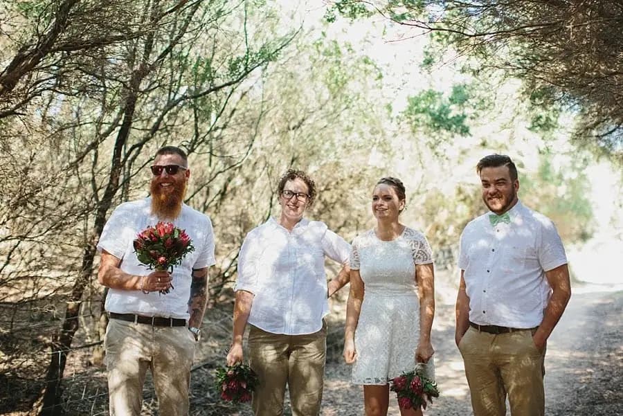 Wedding party walking beneath leafy trees with flower bouquets.