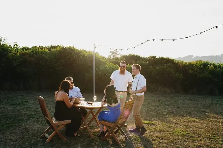 Guests seated and standing around an outdoor table beneath hanging lights.