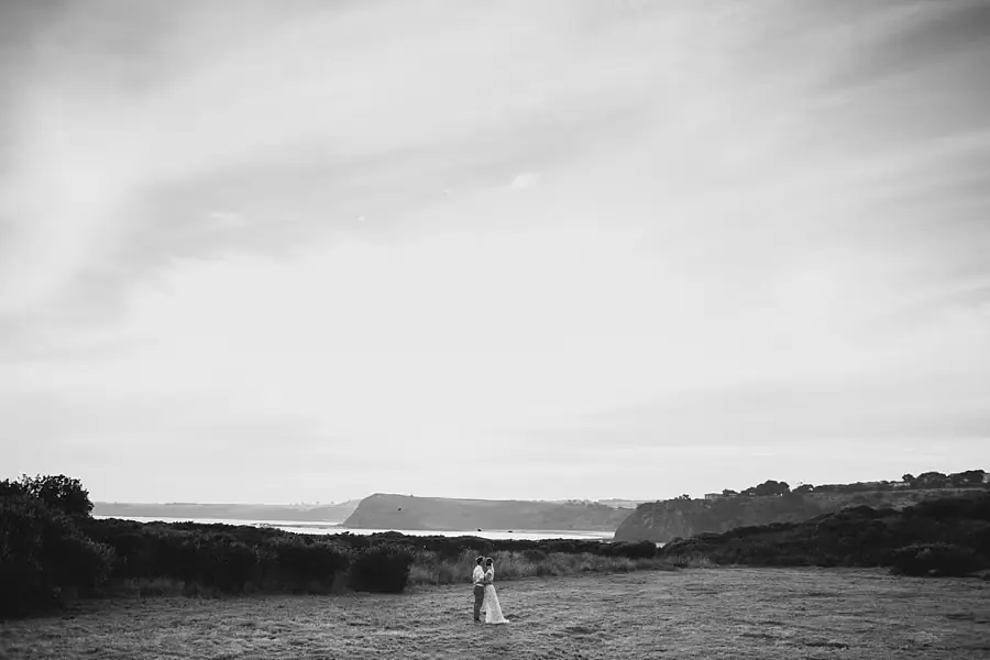 Black-and-white wide view of the couple standing on coastal grass beneath a large sky.