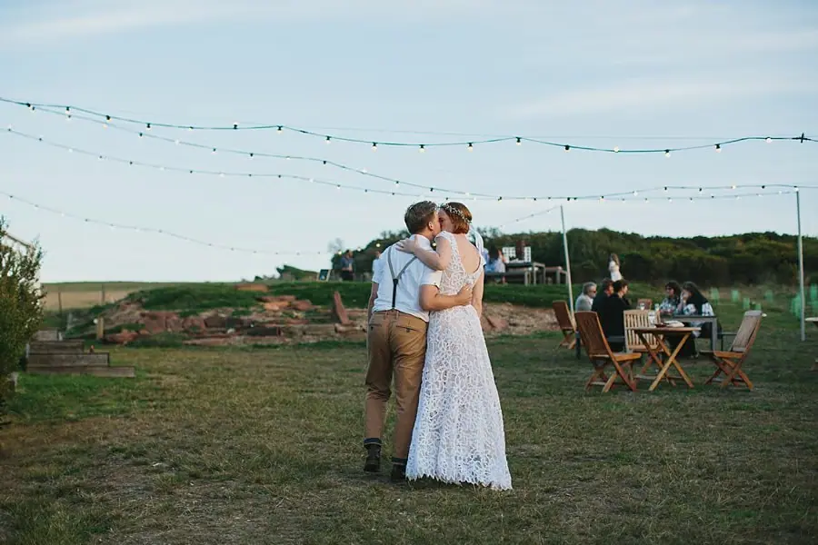 Bride and groom embracing beneath strings of lights on a lawn.
