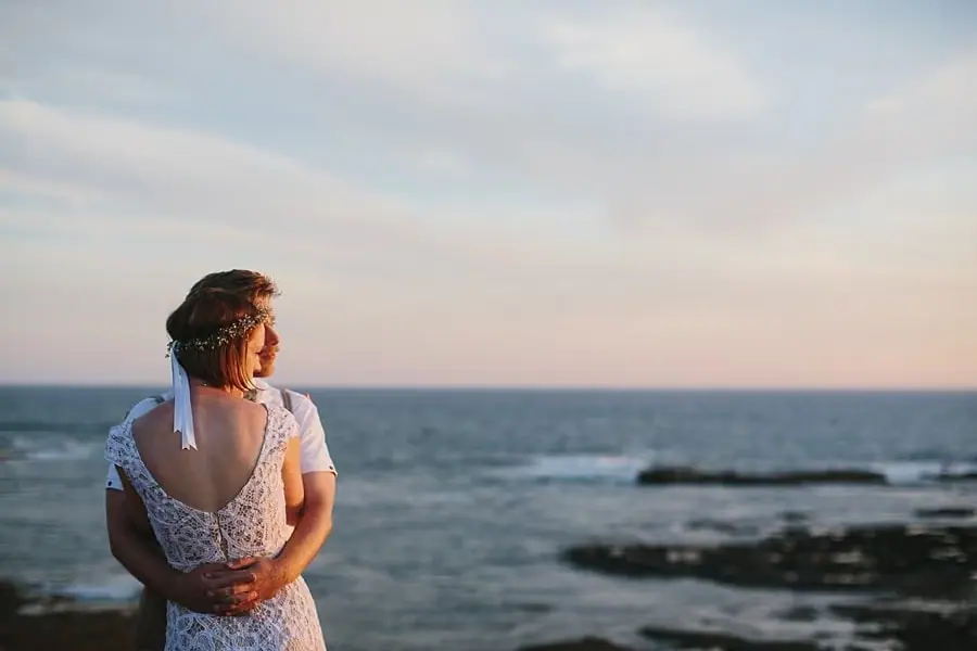 Bride looking across the sea while the groom embraces her from behind.