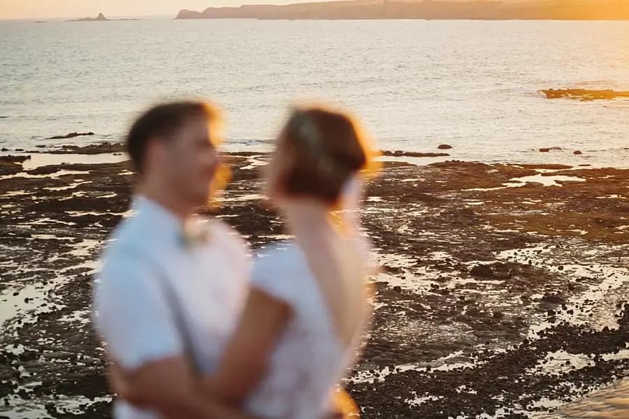 Soft-focus photograph of the couple embracing above a rocky shoreline at sunset.