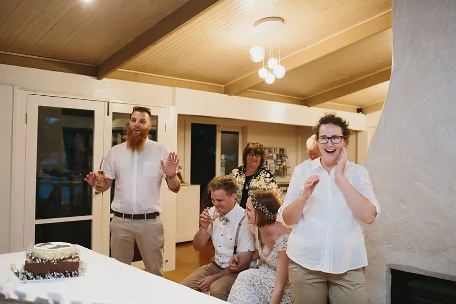 Wedding party laughing around a table with a cake indoors.