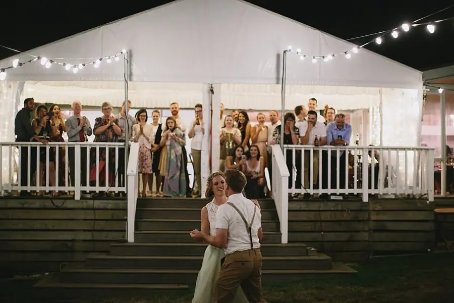 Bride and groom dancing below a veranda crowded with watching guests.