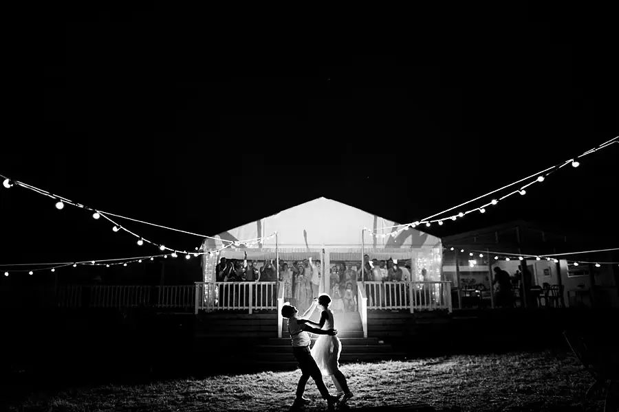 Black-and-white wide photograph of the couple dancing outside an illuminated reception marquee.
