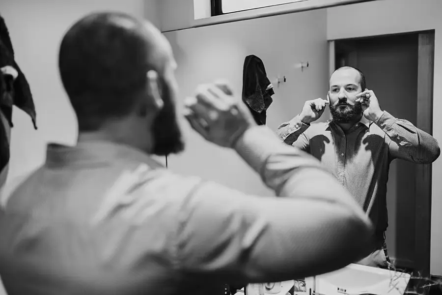 Black and white photograph of a man adjusting his bow tie in a mirror.