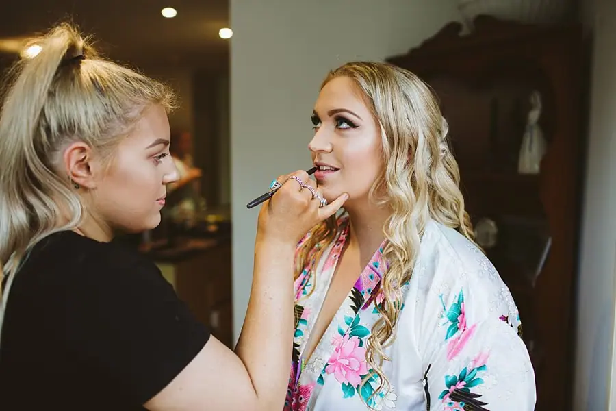 Makeup artist applies lipstick to a woman in a floral robe.