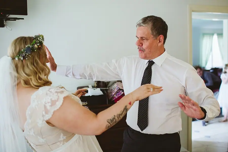 Bride and a man in a shirt reach toward each other indoors.