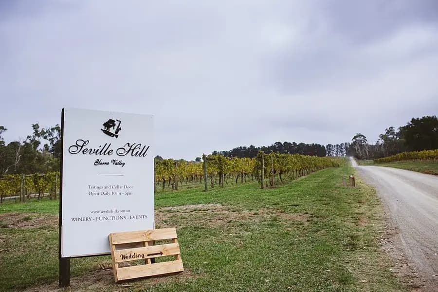 Roadside venue sign beside vineyard rows under grey skies.