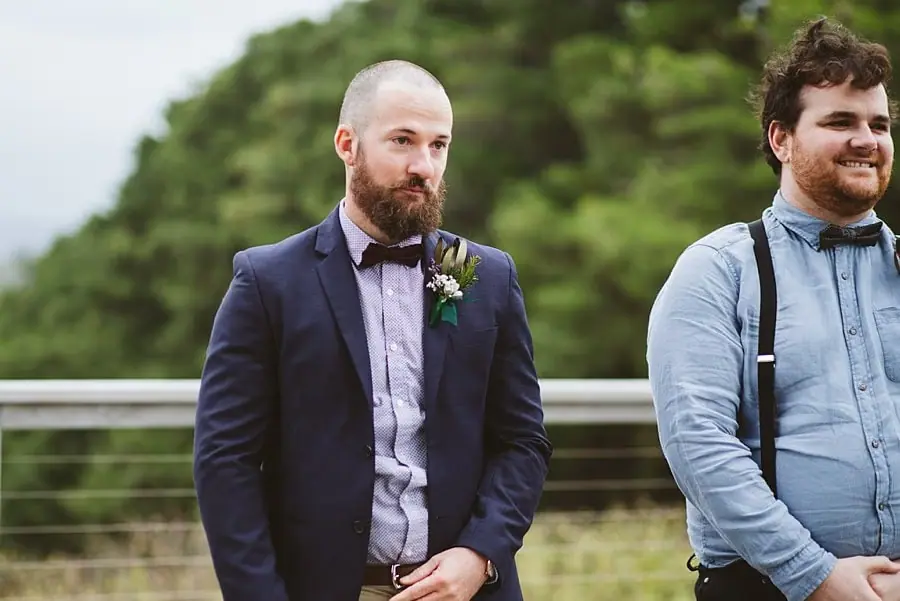 Groom waits with his hands clasped beside a smiling groomsman.